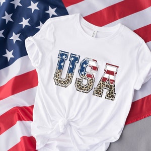 May include: White t-shirt with the word "USA" printed on the front. The letters are in a distressed style and are decorated with an American flag and leopard print.