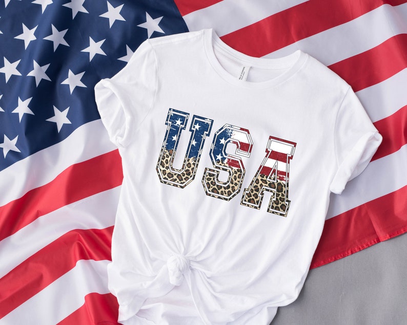 May include: White t-shirt with the word "USA" printed on the front. The letters are in a distressed style and are decorated with an American flag and leopard print.