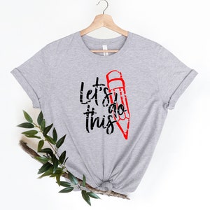 K&ouml;nnte beinhalten: Hellgraues T-Shirt mit dem Text "Let's do this" in Schwarz und einer roten Bleistiftgrafik. Das Shirt h&auml;ngt an einem Holzb&uuml;gel vor wei&szlig;em Hintergrund. Ein Zweig mit gr&uuml;nen Bl&auml;ttern befindet sich in der unteren linken Ecke.