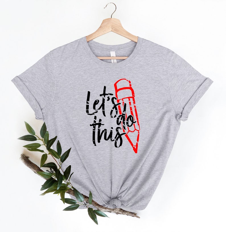 K&ouml;nnte beinhalten: Hellgraues T-Shirt mit dem Text "Let's do this" in Schwarz und einer roten Bleistiftgrafik. Das Shirt h&auml;ngt an einem Holzb&uuml;gel vor wei&szlig;em Hintergrund. Ein Zweig mit gr&uuml;nen Bl&auml;ttern befindet sich in der unteren linken Ecke.