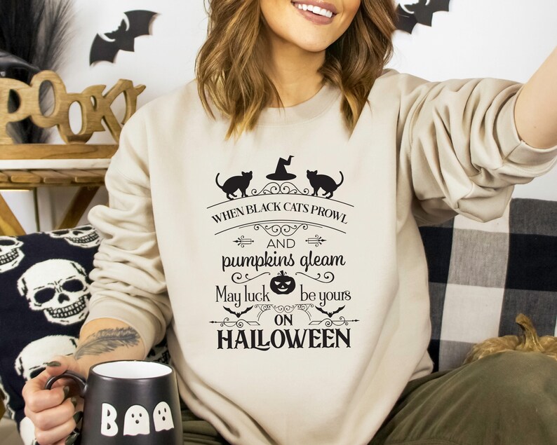 Peut inclure: Un sweat-shirt beige avec un imprim&eacute; graphique noir qui dit "When black cats prowl and pumpkins gleam, may luck be yours on Halloween". Le graphique repr&eacute;sente deux chats noirs, un chapeau de sorci&egrave;re et une citrouille.