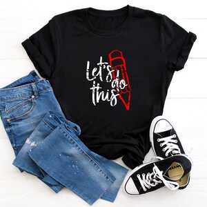 K&ouml;nnte beinhalten: Schwarzes T-Shirt mit dem wei&szlig;en Text "Let's do this" und einer roten Bleistiftgrafik. Das Shirt wird mit blauen Jeans und schwarzen Canvas-Sneakern mit wei&szlig;en Sohlen und Schn&uuml;rsenkeln kombiniert.