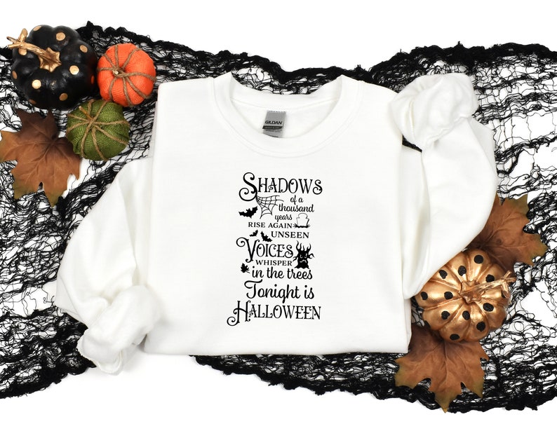 K&ouml;nnte beinhalten: Wei&szlig;es Sweatshirt mit einem schwarzen grafischen Aufdruck, der "Shadows of a thousand years rise again, unseen voices whisper in the trees, Tonight is Halloween" lautet.