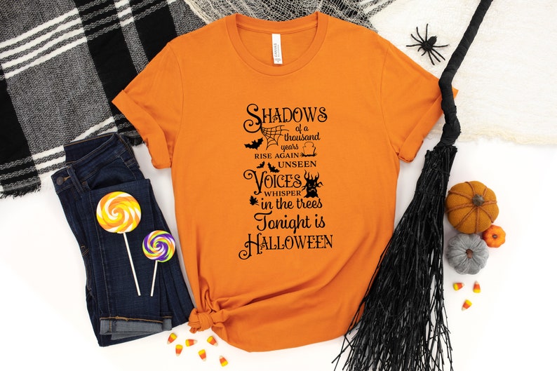 K&ouml;nnte beinhalten: Orangefarbenes T-Shirt mit schwarzem Text, der "Shadows of a thousand years rise again unseen Voices whisper in the trees Tonight is HALLOWEEN" lautet.