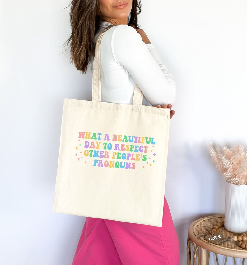 May include: A beige tote bag with the text "WHAT A BEAUTIFUL DAY TO RESPECT OTHER PEOPLE'S PRONOUNS" printed in rainbow colors. The text is in a retro font style.