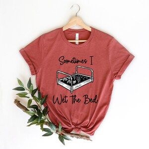 K&ouml;nnte beinhalten: Ein rostfarbenes T-Shirt mit einer schwarzen Grafik eines Bettes, in dem Pflanzen wachsen. Der Text "Sometimes I Wet the Bed" ist unter der Grafik gedruckt.
