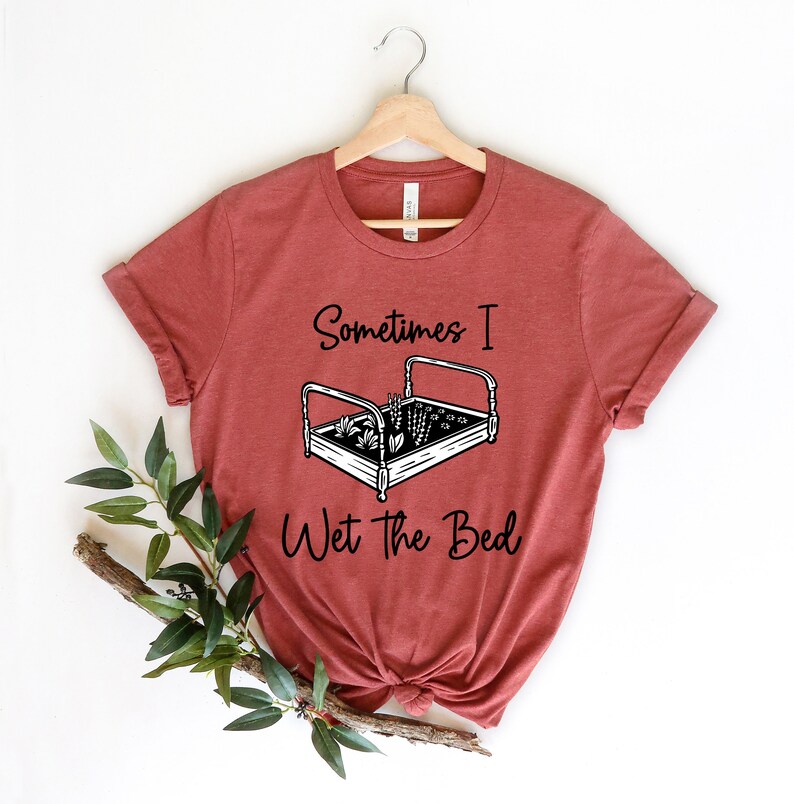 K&ouml;nnte beinhalten: Ein rostfarbenes T-Shirt mit einer schwarzen Grafik eines Bettes, in dem Pflanzen wachsen. Der Text "Sometimes I Wet the Bed" ist unter der Grafik gedruckt.