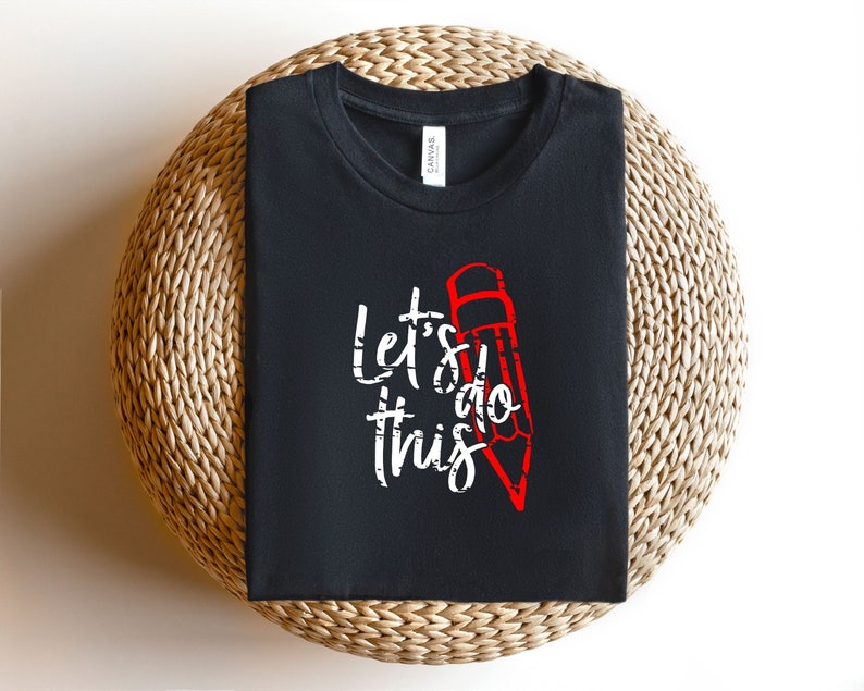 K&ouml;nnte beinhalten: Schwarzes Rundhals-T-Shirt mit dem Schriftzug "Let's do this" in wei&szlig;er Schrift und einer roten Bleistiftgrafik. Das T-Shirt liegt auf einer runden, gewebten Oberfl&auml;che.