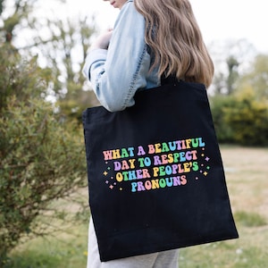 May include: A black tote bag with the text "What a beautiful day to respect other people's pronouns" in rainbow colors. The text is surrounded by small white stars.