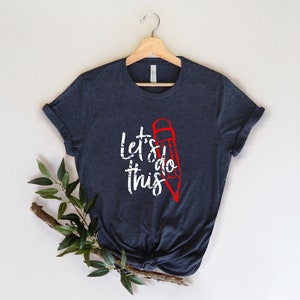 K&ouml;nnte beinhalten: Ein marineblaues T-Shirt mit dem wei&szlig;en Schriftzug "Let's do this", erg&auml;nzt durch eine rote Bleistiftillustration. Das T-Shirt wird auf einem Holzb&uuml;gel vor neutralem Hintergrund pr&auml;sentiert, mit gr&uuml;nen Bl&auml;ttern in der Ecke.