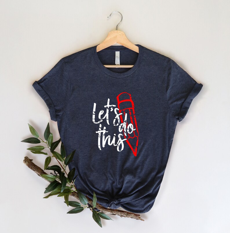 K&ouml;nnte beinhalten: Ein marineblaues T-Shirt mit dem wei&szlig;en Schriftzug "Let's do this", erg&auml;nzt durch eine rote Bleistiftillustration. Das T-Shirt wird auf einem Holzb&uuml;gel vor neutralem Hintergrund pr&auml;sentiert, mit gr&uuml;nen Bl&auml;ttern in der Ecke.