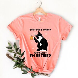 以下が含まれることがあります： コーヒーカップを持った白黒の猫が描かれたピンクのTシャツ。Tシャツには「今日は何曜日？誰が気にする？私は引退した。」という文字が書かれています。
