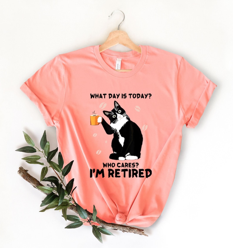 以下が含まれることがあります： コーヒーカップを持った白黒の猫が描かれたピンクのTシャツ。Tシャツには「今日は何曜日？誰が気にする？私は引退した。」という文字が書かれています。