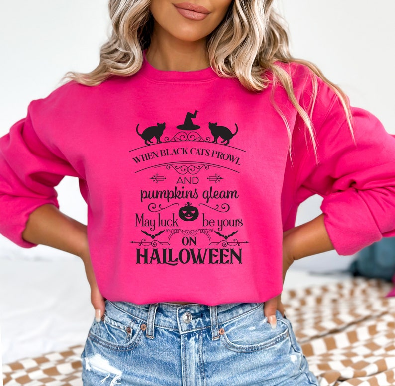 Peut inclure: Un sweat-shirt rose avec un imprim&eacute; graphique noir qui dit "When black cats prowl and pumpkins gleam, may luck be yours on Halloween". Le graphique comprend &eacute;galement un chat noir, un chapeau de sorci&egrave;re, une citrouille et des chauves-souris.