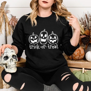Może przedstawiać: Czarna bluza z trzema wyrzeźbionymi twarzami dyni i białym napisem "trick-or-treat". Bluza jest częścią kolekcji o tematyce Halloween. Obraz zawiera r&oacute;wnież dekoracyjną czaszkę i dynie.
