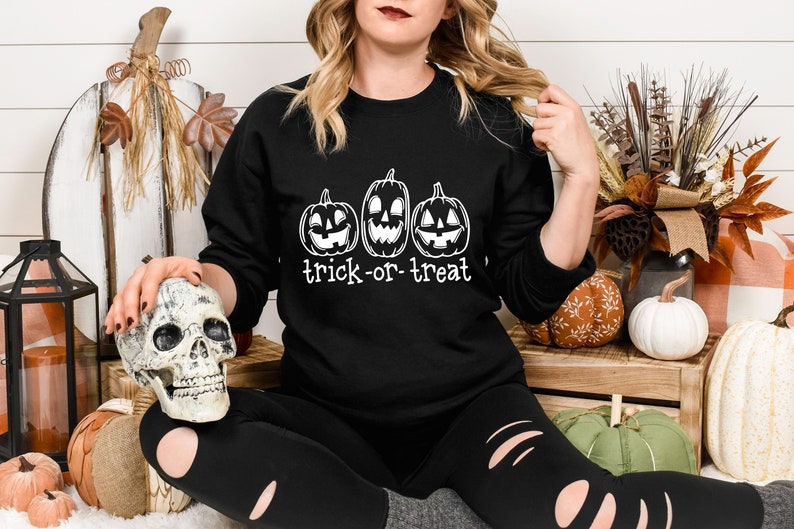 Może przedstawiać: Czarna bluza z trzema wyrzeźbionymi twarzami dyni i białym napisem "trick-or-treat". Bluza jest częścią kolekcji o tematyce Halloween. Obraz zawiera r&oacute;wnież dekoracyjną czaszkę i dynie.