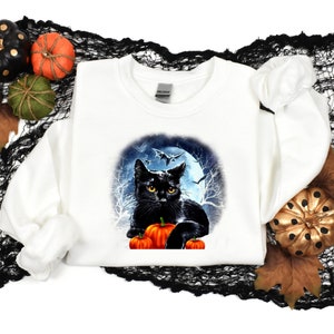 May include: White sweatshirt with a black cat and pumpkins in front of a full moon and bats. The cat is looking at the viewer.