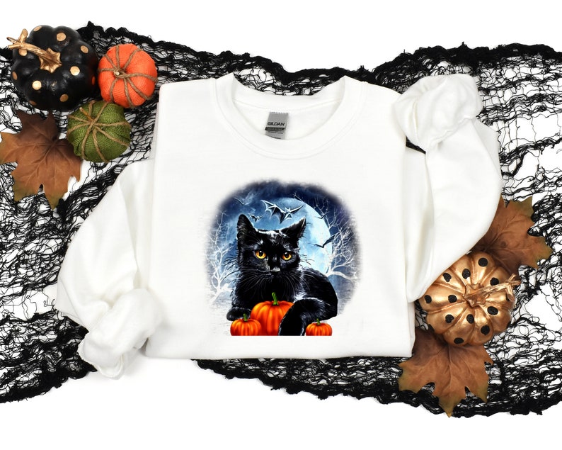 May include: White sweatshirt with a black cat and pumpkins in front of a full moon and bats. The cat is looking at the viewer.