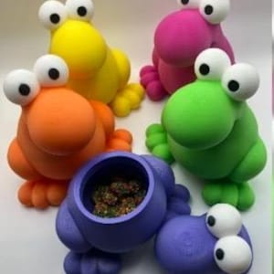 Puede incluir: Un conjunto de recipientes coloridos y caprichosos con forma de rana. Las ranas son de color amarillo, naranja, rosa, verde y morado. Una rana est&aacute; abierta, revelando un interior oscuro. Cada rana tiene ojos grandes y blancos y pies peque&ntilde;os.