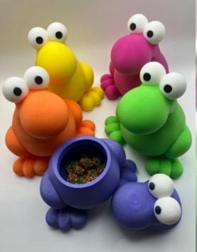 Puede incluir: Un conjunto de recipientes coloridos y caprichosos con forma de rana. Las ranas son de color amarillo, naranja, rosa, verde y morado. Una rana est&aacute; abierta, revelando un interior oscuro. Cada rana tiene ojos grandes y blancos y pies peque&ntilde;os.