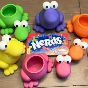 Puede incluir: Una colecci&oacute;n de recipientes coloridos con forma de rana en verde, morado, naranja, amarillo y rosa, cada uno con ojos grandes y saltones. Tambi&eacute;n hay una caja de caramelos Nerds Rainbow. Los recipientes est&aacute;n colocados sobre una superficie de madera.