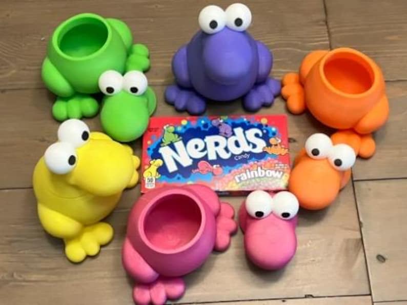 Puede incluir: Una colecci&oacute;n de recipientes coloridos con forma de rana en verde, morado, naranja, amarillo y rosa, cada uno con ojos grandes y saltones. Tambi&eacute;n hay una caja de caramelos Nerds Rainbow. Los recipientes est&aacute;n colocados sobre una superficie de madera.
