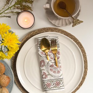 May include: A white ceramic plate with a gold fork and spoon wrapped in a white napkin with pink floral print. The plate is on a brown woven placemat.