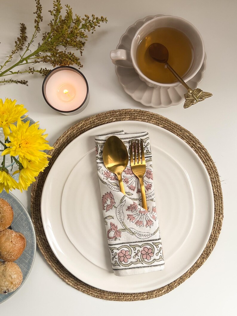 May include: A white ceramic plate with a gold fork and spoon wrapped in a white napkin with pink floral print. The plate is on a brown woven placemat.