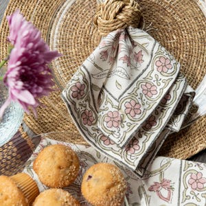 May include: A set of four muffins on a white plate with a floral pattern. The muffins are topped with sugar crystals. A napkin ring made of rope is holding a white napkin with a pink and green floral pattern.