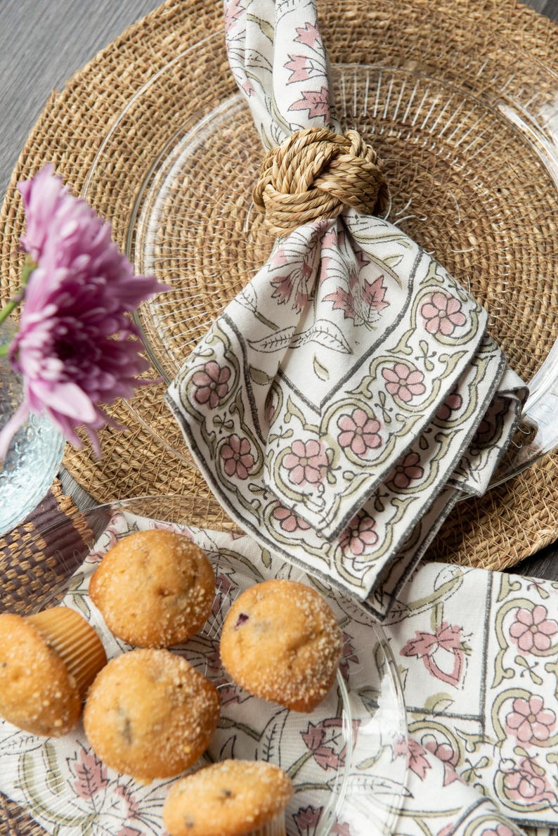 May include: A set of four muffins on a white plate with a floral pattern. The muffins are topped with sugar crystals. A napkin ring made of rope is holding a white napkin with a pink and green floral pattern.