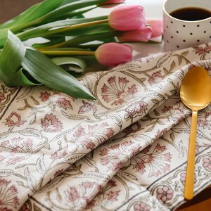 May include: A white cotton tea towel with a pink floral pattern and a gold spoon on top. The towel is folded in half and has a border of green and pink.