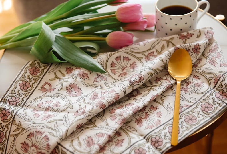 May include: A white cotton tea towel with a pink floral pattern and a gold spoon on top. The towel is folded in half and has a border of green and pink.