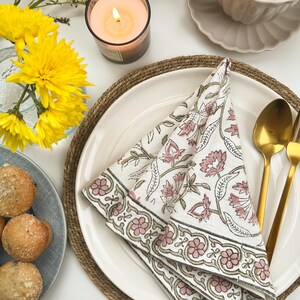 May include: A white cotton napkin with a pink and green floral print is folded and placed on a white plate with a brown woven placemat. A gold fork and spoon are placed on the plate.