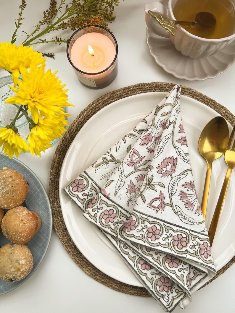 May include: A white cotton napkin with a pink and green floral print is folded and placed on a white plate with a brown woven placemat. A gold fork and spoon are placed on the plate.