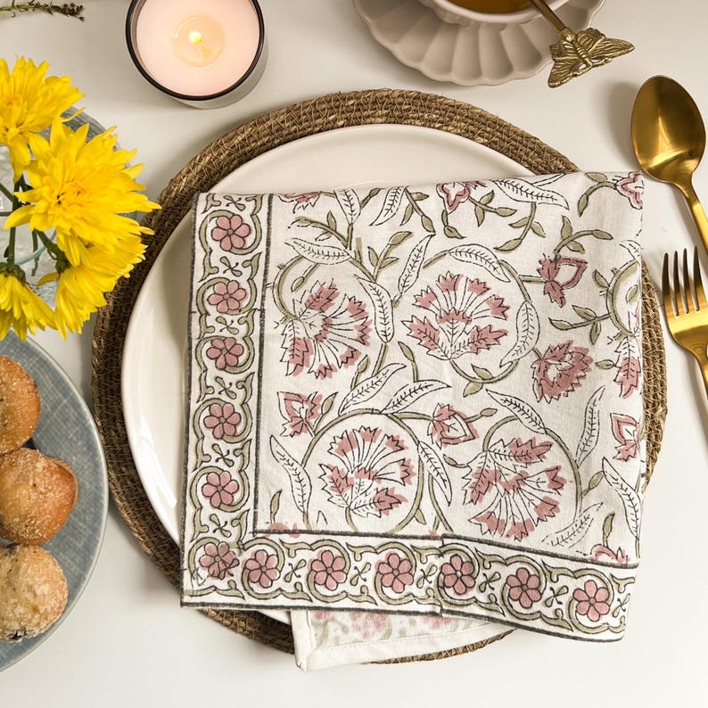 May include: A white cotton napkin with a pink and green floral print. The napkin is folded and placed on a woven placemat.