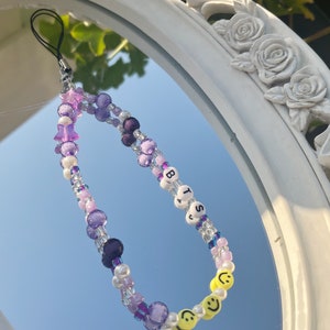 May include: A purple, white, and yellow beaded phone charm with the letters "BTS" and smiley faces.