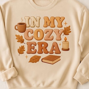 以下が含まれることがあります： クリーム色のスウェットシャツ。茶色とオレンジ色の文字で「IN MY COZY ERA」と書かれています。デザインには、コーヒーカップ、葉、ろうそく、本が含まれています。すべて暖かみのある秋の色です。クルーネックと長袖です。