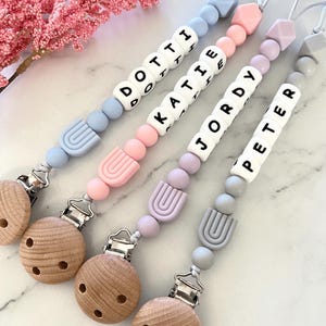 Op de afbeelding: Vier gepersonaliseerde baby fopspeenclips in pastelkleuren. Elke clip heeft een houten ronde clip, siliconen kralen en een naam gespeld in witte letterkralen: Dottie, Katie, Jordy en Peter. Een roze bloementak staat op de achtergrond.