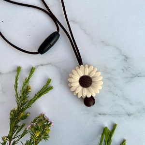 May include: A white daisy flower charm necklace with a brown cord and a black plastic clasp. The necklace is on a white marble background.