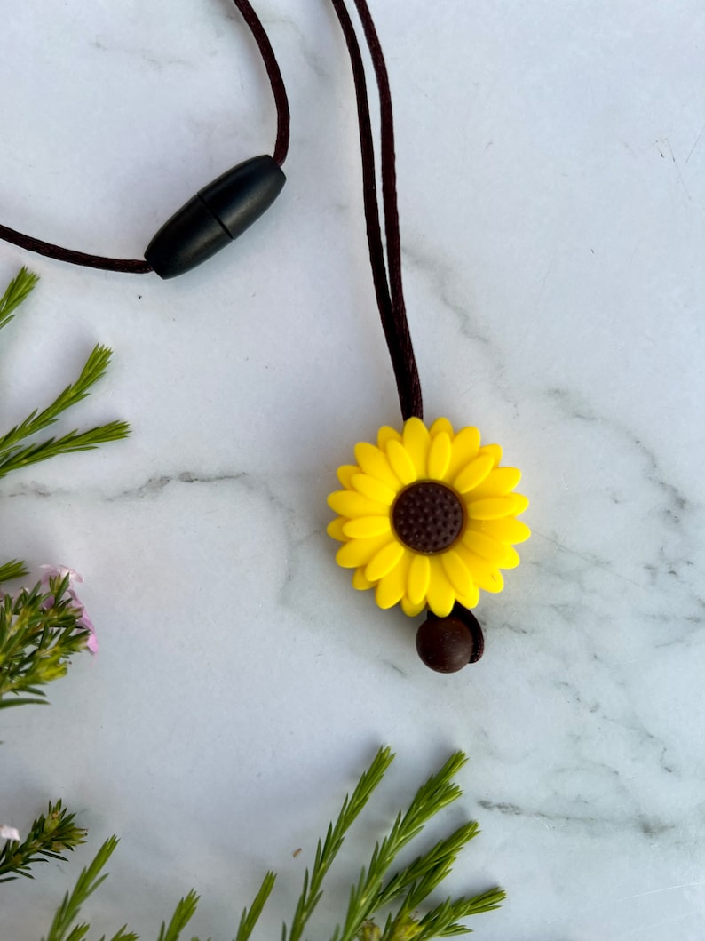 May include: A yellow sunflower silicone necklace with a brown center and a brown bead on the cord. The necklace is on a white marble surface with green foliage.