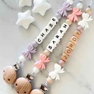 May include: Three personalised dummy clips with names spelled out in letter beads. Each clip features a wooden dummy holder, bow accents, and various bead colours. Three white star-shaped toys are also present.