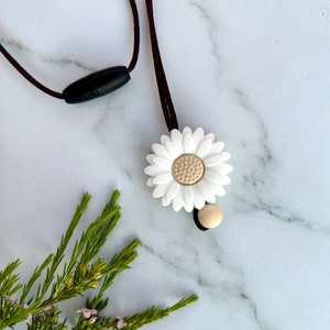 May include: A white daisy charm necklace with a brown cord and a black plastic clasp. The charm has a gold center and is attached to the cord with a small brown bead.