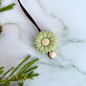 May include: A green daisy charm with a white center on a brown cord. The charm is attached to a small, beige bead.