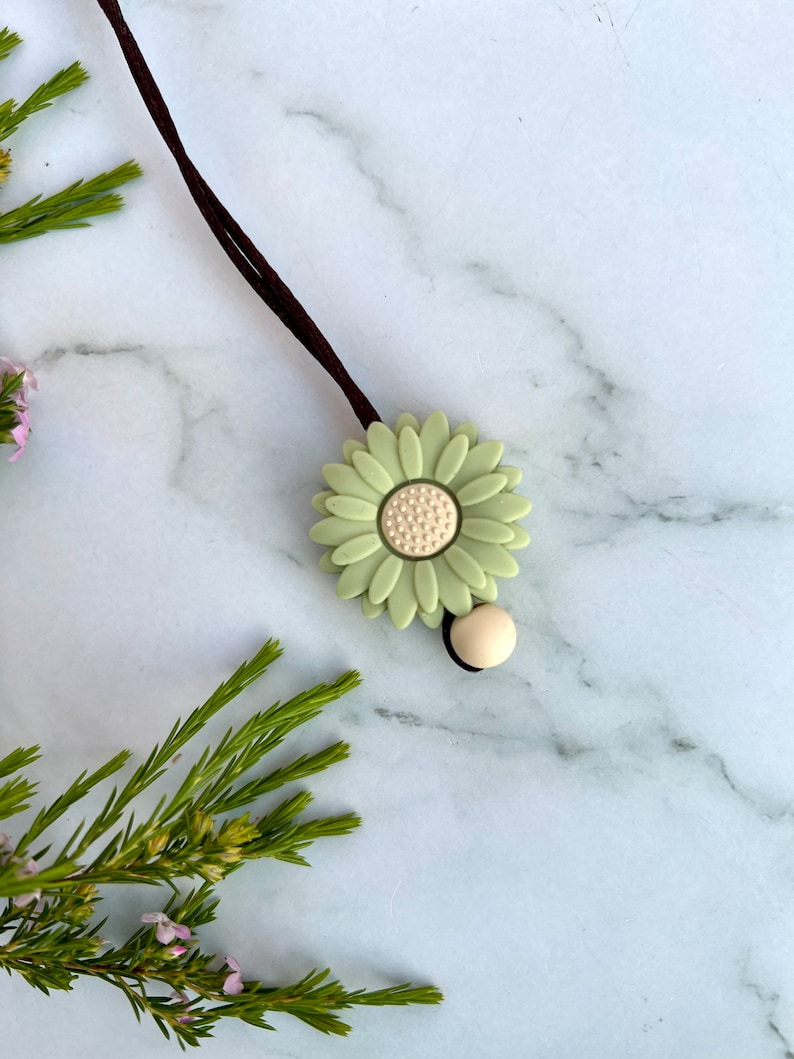 May include: A green daisy charm with a white center on a brown cord. The charm is attached to a small, beige bead.