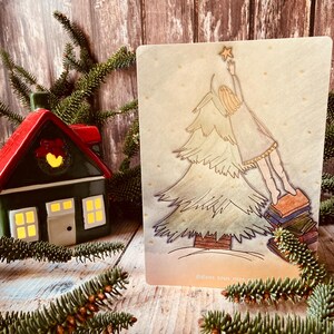 Könnte beinhalten: Weihnachtskarte mit einer handgezeichneten Illustration einer Person, die nach einem Stern über einem Weihnachtsbaum greift. Die Karte steht vor einem Holz Hintergrund, mit einem kleinen Haus und Tannenzweigen im Vordergrund, die eine saisonale Szene schaffen.