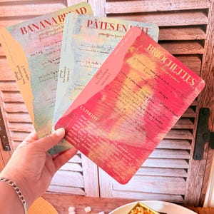 Peut inclure: Une main tient trois cartes de recettes avec des designs colorés et du texte manuscrit. Les cartes présentent des titres tels que "Banana Bread", "Pates Ente" et "Briochettes". L'arrière-plan comprend un volet en bois et une assiette avec de la nourriture.