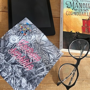 May include: A black and white patterned microfiber cleaning cloth with the words "Lettice & Sons" printed in pink. The cloth is lying on a wooden surface next to a book titled "Un Manoir en Cornouailles" by Eve Chase, a black e-reader, and a pair of black framed glasses.