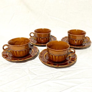 Può includere: Set di quattro tazze da tè e piattini in ceramica marrone. Ogni tazza ha un corpo arrotondato con un design a coste e un manico curvo. I piattini hanno un bordo smerlato. Il set è esposto su una superficie bianca.