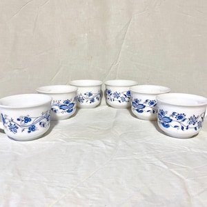 Könnte beinhalten: Sechs weiße Keramikbecher mit blauem Blumendesign. Die Becher haben eine abgerundete Form und einen Henkel. Das blaue Blumenmuster umschließt die Becher. Die Becher sind auf einer weißen Oberfläche angeordnet.