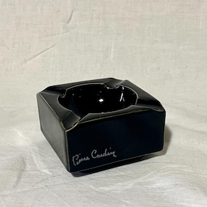 Può includere: Un posacenere quadrato nero con finitura lucida. Il posacenere ha un centro incassato e lati angolati. Le parole "Pierre Cardin" sono scritte in corsivo sul lato.